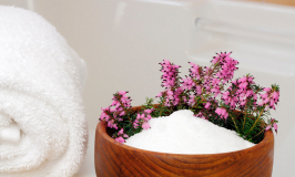 White bath towel on side of tub next to wooden bowl containing epsom salt and pink flowers.